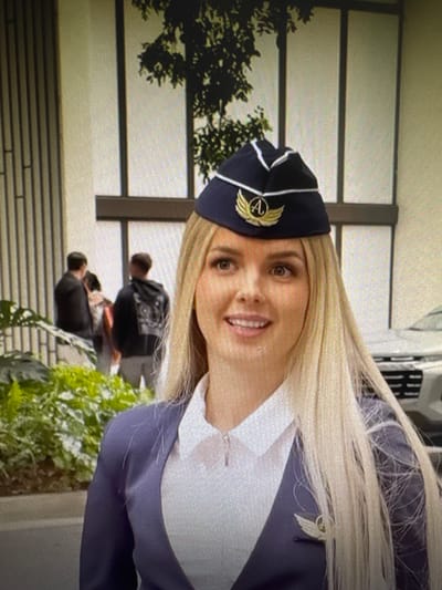 The image features a close-up of a woman dressed in a flight attendant uniform, including a dark blue hat with a gold emblem and a white shirt. She has blonde hair and is smiling at the camera. The background is blurred, but appears to be an outdoor setting with modern architecture. In the background, two other individuals can be seen from behind, wearing dark clothing. The lighting suggests it is daytime. The overall mood is cheerful and professional. The image is oriented sideways.