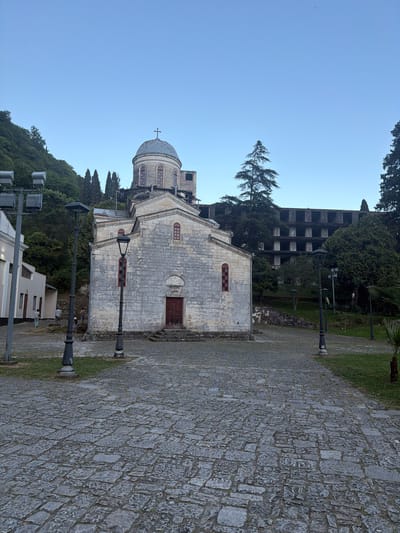 The image shows a view of the New Athos Monastery in New Athos, Abkhazia. The monastery is a prominent religious building with a distinctive dome topped with a cross. The building is constructed of stone and features several arched windows with red frames. In front of the monastery is a paved courtyard made of cobblestones. Several lampposts are present in the courtyard. To the left of the monastery is a white building. In the background, a steep, tree-covered hill rises behind the monastery. Further in the background and to the right, a large, multi-story concrete structure, appearing somewhat derelict, is visible amidst the trees. The sky is clear and blue. The image appears to have been taken during daylight hours, likely in the morning or late afternoon due to the soft lighting. A few individuals are visible in the distance, walking in the vicinity of the white building.