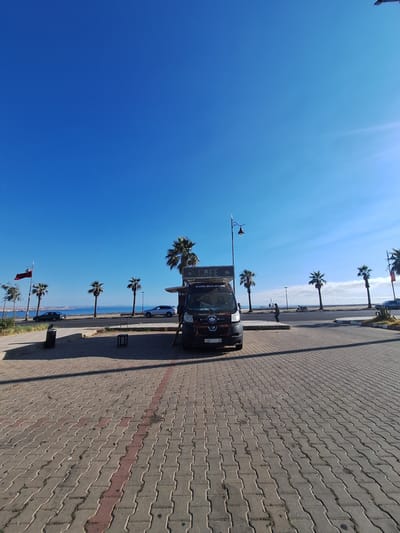 The image depicts a sunny day in Safi, Morocco, with a clear blue sky and bright sunlight casting long shadows. The scene is a waterfront promenade paved with interlocking bricks, some of which are reddish-brown, creating a decorative line. Along the promenade, a row of palm trees stands tall, their fronds catching the light.
In the foreground, a black van, possibly a food truck, is parked. It has signage on its side, including text that reads "CAFÉ" and "Café des amies". The van also has a license plate that appears to be "23481 | 55". A street lamp is attached to the side of the van. Further down the promenade, other vehicles are parked, and a few people are visible walking or standing. One person appears to be near a trash bin.
Beyond the promenade, the calm blue sea stretches out to the horizon. A red flag is visible on a flagpole among the palm trees towards the lower part of the image. The overall impression is one of a pleasant, leisurely day at the coast. The time of day is clearly daytime, given the strong sunlight and pronounced shadows, and the weather is fair and sunny.