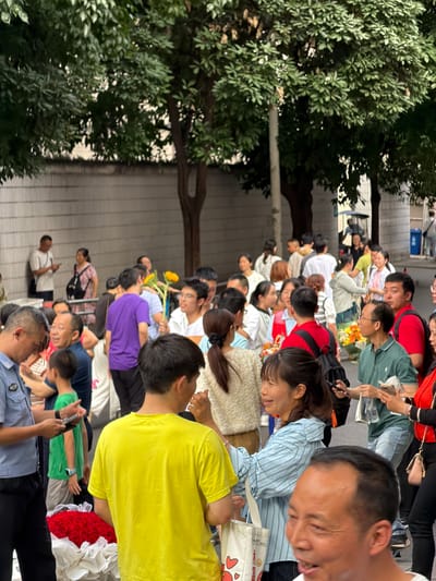 A dense crowd of people, predominantly East Asian, are gathered on a paved street in front of a concrete wall, possibly on the outskirts of a city. Several individuals are holding bouquets of flowers, notably a large arrangement of red roses at the forefront. Some people are looking at phones, while others are engaged in conversation. The presence of trees suggests an outdoor urban environment. The individuals appear to be of various ages and genders, dressed in casual clothing. The overall atmosphere seems to be one of a public gathering or event.