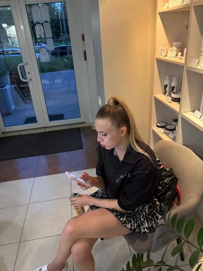 In Romanovka, Russia, an indoor scene depicts a young woman seated on a light grey upholstered chair, positioned to the right of a large white-framed glass double door. The woman, with blonde hair tied in a high ponytail, wears a black collared shirt with partially visible white lettering ("LUMOUS") on the left chest and a leopard print ruffled skirt. She has red nail polish on her fingers and white open-toed sandals on her feet, with her legs crossed at the knees. Her gaze is directed downwards at a pink smartphone held in her left hand, while her right hand holds a golden-yellow beverage can. A black plastic bag is draped over the back of her chair, partially obscured.
The interior features a tiled floor composed of light grey/white sections in the foreground and darker reddish-brown sections with lighter veins closer to the door. The wall to the left is plain beige, while the wall to the right features light-colored shelving units displaying various cosmetic or personal care products in white and black containers, some with visible price tags. A small green plant is situated on the lower shelf, and a larger green leafy plant (possibly a ZZ plant) occupies the lower right foreground.
Through the glass double door, the exterior is visible, showing a dark paved area, green grass, and trees under what appears to be evening or twilight lighting. A silver sedan and a dark-colored SUV are discernible in the distance. Reversed text, including "ВАНИЛА" and "BRANIMANHA", is visible on the glass due to reflections or interior signage. Several opaque white, irregularly shaped stickers are affixed to the glass panels. A dark grey doormat rests immediately inside the door.