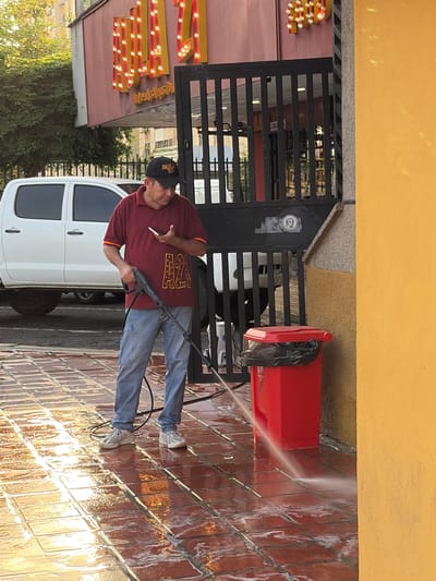 This image, set in Guayana City, Venezuela, depicts a man actively engaged in pressure washing a tiled ground surface. The scene appears to be an outdoor area, possibly a business entrance or patio, bordered by a black metal gate and a yellow building wall.
The central subject is a man, likely middle-aged, wearing a maroon short-sleeved polo shirt with a gold "AZ" or "A7" design on the front, blue jeans, and light-colored sneakers. He also wears a dark baseball cap. In his right hand, he holds a black and blue pressure washer gun, directing a stream of water onto the reddish-brown tiled floor, which is visibly wet and reflective. A white cigarette is held between the fingers of his left hand.
Behind the man, a white pickup truck is partially visible. To his right, behind the gate, stands a bright red trash can with a black garbage bag liner. On the far left, a sign adorned with yellow and red exposed light bulbs is visible, and some indistinct gold text can be faintly seen on it, possibly reading "FRESH" or a similar word, along with what might be "100%." The ground is a grid of square, reddish-brown tiles, glistening from the water. Green foliage and a building with a pink wall are seen in the background on the left.
The lighting suggests it is daytime, with sunlight reflecting off the wet surfaces. The man appears focused on his task. A small circular sticker with an unclear logo or face is affixed to the black gate. The activity indicates maintenance or cleaning work is underway.