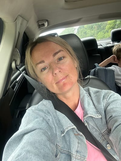 An adult female with blonde hair, wearing a pink t-shirt and a denim jacket, is seated in the driver's seat of a car, looking directly at the camera. Her seatbelt is fastened. Her face shows a slightly flushed complexion on her cheeks and chin, and her lips appear shiny. A child with light brown hair is visible in the backseat behind her, wearing a white t-shirt and leaning against the car's headrest. A blue backpack is situated between them. The interior of the car, including black leather seats and a gray headliner, is visible. The exterior through the rear window shows greenery and a cloudy sky. The image is a selfie taken from within the vehicle.