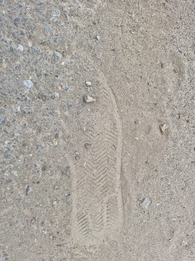 The image displays a close-up, overhead view of a sandy surface with embedded small stones and gravel. A distinct shoe print, characterized by a herringbone pattern, is clearly visible pressed into the sand, oriented diagonally from the lower left to the upper right of the frame. The lighting suggests an outdoor environment. There is no indication of the city of Nyagan, Russia, or any specific location within a city. The scene is devoid of people or any other discernible subjects or objects beyond the natural elements of the ground and the imprint.