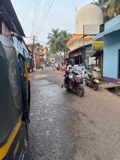 The image displays a street scene within Gokarna, India, characterized by urban infrastructure and local activity under a hazy sky.

In the immediate foreground on the left, the rear and side portion of a yellow and black auto-rickshaw is visible, oriented parallel to the direction of the street. The road surface is asphalt, exhibiting wear, exposed aggregate, and several small, dark, irregularly shaped wet patches or shallow puddles.

In the midground, a maroon-colored Honda Activa scooter is in motion, occupied by three individuals. A male driver, wearing a dark helmet, operates the scooter. Directly behind him, a female passenger in a dark top and light-colored trousers sits sideways. A third individual, partially obscured by the female passenger, is positioned behind her. To the right of this moving scooter, a light gold-colored Honda Activa scooter is parked facing away from the viewer, positioned on the shoulder of the road.

The street is lined with multi-story buildings on both sides. On the left, several structures function as shops, displaying assorted textiles and clothing hanging from their facades. A dense network of black electrical or communication wires spans overhead across the street. Further down the street, several pedestrians are visible walking.

On the right side of the street, a prominent white, dome-shaped structure rises above the surrounding buildings, featuring a red and white trident-like symbol at its apex. This structure is indicative of a temple, consistent with the Mahabaleshwar Temple complex in Gokarna. Directly below this dome, a multi-story building hosts various businesses, as indicated by signs. Visible signs include "Prema Dental Clinic and Maxillofacial Surgical Centre" and "Gokarna Credit Co-Operative Society," with text in both English and Kannada scripts. Adjacent to a light blue building on the far right, a brown dog lies sleeping on the dirt curb. Multiple palm trees are interspersed among the buildings and in the background.