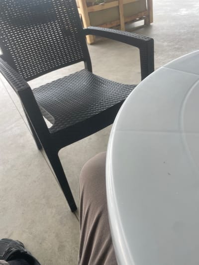 The image shows an outdoor seating area with a black plastic chair and a white round table. A person's legs, clad in brown pants, are visible in the foreground, seated at the table. The ground is concrete, and in the background, there is a wooden structure that appears to be part of a cafe or restaurant. The overall scene suggests a casual outdoor dining or relaxation setting in Malevo, Bulgaria.