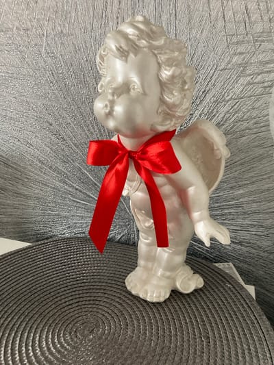 A white, pearlized figurine of a cherub, adorned with a red satin ribbon tied in a bow around its neck, stands on a textured, dark gray circular placemat. The cherub has curly hair and plump cheeks. Behind the figurine is a shimmering, textured silver background that resembles a fan or sunburst. The figurine is positioned centrally in the frame, with its body slightly turned to the right. The lighting appears to be soft, highlighting the pearlescent finish of the cherub. The image does not provide any information that would allow for localization to Balabanovo, Russia.