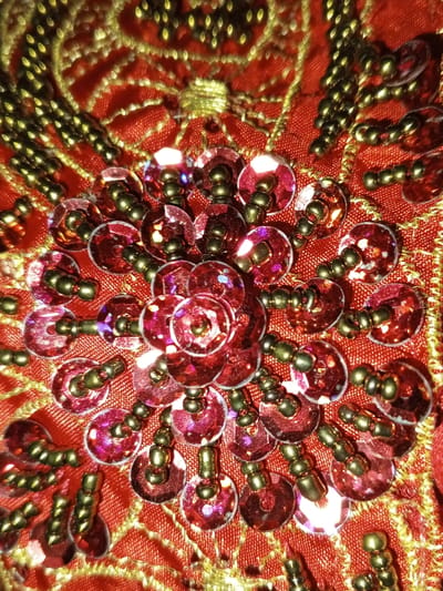 This image presents a detailed, close-up view of an elaborately decorated textile from Keo Kouang, Laos. The foundation is a textured red fabric, over which complex patterns of gold thread embroidery are visible, forming intertwined lines and floral-like motifs. Scattered across and largely overlaying the gold embroidery are numerous circular, translucent red sequins, each approximately 3-5 millimeters in diameter, exhibiting iridescent properties with flashes of purple and pink under direct light. Each sequin is individually secured to the fabric by a cluster of small, dark gold-toned spherical beads, which are threaded through the central hole of the sequin, creating a slightly raised attachment point. The sequins are arranged in a dense central cluster, resembling a floral bloom, with additional sequins and beaded attachments extending outwards across the red fabric and gold embroidery. No people or external environment are depicted; the scene focuses entirely on the textile's surface.