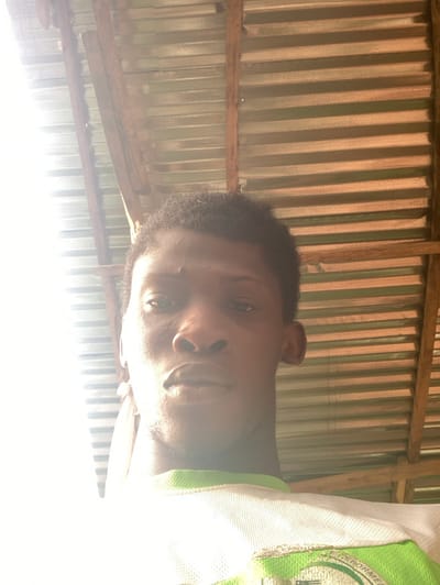 The image displays a young adult male located in Agege, Nigeria. The subject has dark skin, short, dark, curly hair, and faint stubble on his chin and jawline. He is wearing a white shirt, possibly mesh fabric, with green trim visible on the collar and upper chest, and a partially discernible green logo. The subject faces directly towards the camera with a neutral expression. A bright light source is positioned to the left, causing significant overexposure on the left side of the subject's face and upper body. The background features a corrugated metal roof structure with horizontal ridges, supported by rough-hewn wooden rafters positioned diagonally and horizontally across the upper portion of the frame.