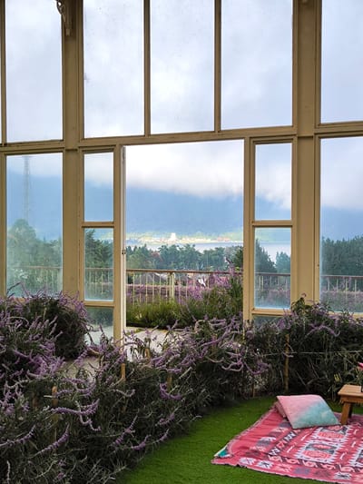 The scene depicts an indoor setting in Candikuning, Indonesia, looking out through a large window toward a lake and mountains on a cloudy day. The window, framed in light brown wood, consists of multiple panes, with the central section forming an open doorway leading onto a balcony with a metal railing.

Inside, a patch of artificial green grass hosts a pink patterned blanket, a turquoise gradient pillow, and a small wooden side table. Purple flowering plants, possibly lavender, fill the space immediately inside the window, creating a foreground border.

Beyond the balcony, the view encompasses a body of water, presumably a lake, surrounded by lush greenery and mountain slopes. In the distance, structures resembling buildings are visible, potentially part of a town or village along the shore. A tall communications tower stands on a hill in the left portion of the scene. The sky is overcast, and the light suggests a cool, possibly damp atmosphere. There are no people present in the photograph.