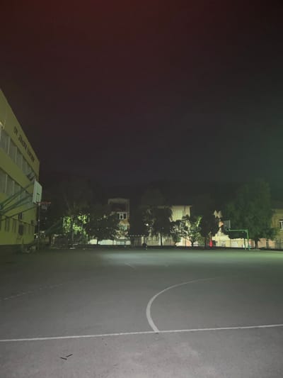 This is a night-time photograph of an outdoor basketball court located in front of a yellow building with the text "OV EMOTIV TECHNER" visible on its facade. The court is marked with white lines, including a semi-circle. A basketball hoop and backboard are positioned to the left of the frame, attached to a green metal support structure. Several trees are scattered around the court, and beyond them, buildings are visible, some with illuminated windows. The sky above is dark with a few faint stars. The scene appears to be situated in a schoolyard or a similar public recreational area. The overall impression is one of a quiet, possibly deserted, educational or sports facility after dark in Dupnitsa, Bulgaria.