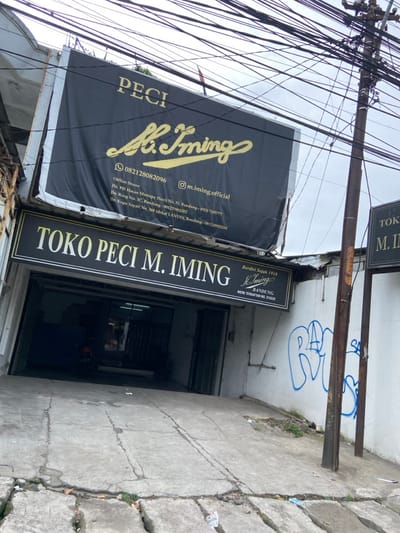 The image shows a storefront with a sign for a hat store called "Toko Peci M. Iming" in Kecamatan Margahayu, Indonesia. The storefront is black with gold lettering and has a large sign above it that reads "Peci M. Iming" in gold lettering. The storefront is located on a street with a telephone pole in front of it. There are multiple power lines running across the top of the image. The storefront is located on an incline and the image shows the concrete path leading up to the storefront with some cracks in it. There is blue graffiti on the wall to the right of the telephone pole.  The shop has a large glass window with a sign that says "Berdiri Sejak 1918 M. Iming Bandung" in gold lettering. There is a car visible in the window of the storefront.  The shop has a phone number of 082128082096 and an Instagram handle of @m.iming_official.  There is a small sign on the wall of the building to the right of the telephone pole that reads "Toko M. Iming".  The sign has faded gold lettering and is painted on a white background.  The environment is urban with a building in the background that is out of focus.  The photo is taken during the day.