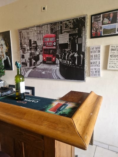 The image shows a wall adorned with various framed pictures and prints. Above is a framed photograph and a wall-mounted print. Below are prints with motivational messages. One depicts a red double-decker bus in a black and white cityscape. Adjacent to the wall is a wooden bar or cabinet with a partially visible bottle of wine on top of it. The background includes a glimpse of what appears to be a floor or tiled surface.