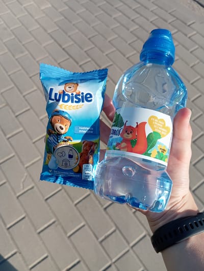 In Radzymin, Poland, a person holds a blue plastic bottle of water and a blue packet of "Lubisie" cookies. The person's hands are visible, with a dark fitness tracker on their wrist. The background consists of grey interlocking paving stones, suggesting an outdoor urban setting. The lighting indicates it is daytime, with shadows present. The "Lubisie" cookie packet features a cartoon bear, while the water bottle label has an illustration of a squirrel. The image focuses on these two items held by the person.