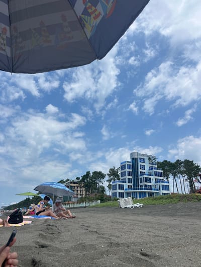 The image depicts a scene on a black sand beach in Shekvetili, Georgia, on a cloudy day. In the foreground, the underside of a large beach umbrella, decorated with images of lighthouses and sailboats, is visible against a bright blue sky with scattered white clouds. Further back on the beach, several people are relaxing under umbrellas. Two women are sitting on towels, with one appearing to be showing something to the other. A black bag is placed on the sand near them. In the middle ground, a modern, multi-story building with blue-tinted windows and balconies stands out against a line of pine trees. Stacked white lounge chairs are visible in front of the building. The ground is covered in dark, granular sand. The overall atmosphere suggests a relaxed beach setting.