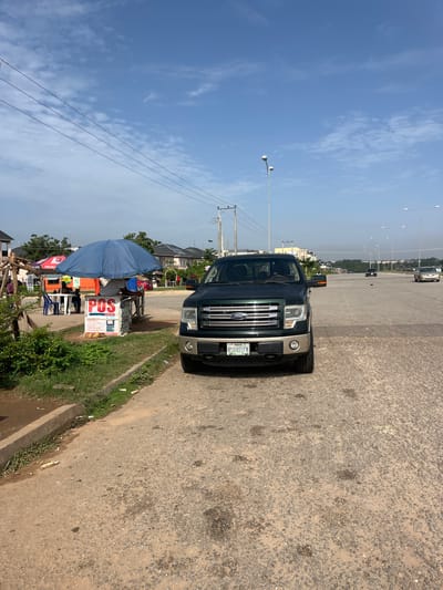 In Karum, Nigeria, a dark green Ford F-150 truck is parked on the right side of a paved road, facing forward. The truck's license plate reads "RSH 871FN". To the left of the truck, a blue umbrella shades a small stand labeled "POS" with various financial services listed. A person is seated behind the POS stand. Behind the stand, there are two orange tents and some tables and chairs. The road itself is wide with multiple lanes and illuminated by streetlights. Further down the road, other vehicles are visible, and residential buildings line the background. The sky is clear with scattered clouds, and the environment appears to be a commercial or roadside area with some greenery on the left.