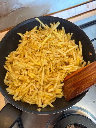 A top-down shot shows a black skillet on a stovetop, filled with fried potatoes and a wooden spatula. The potatoes are cut into thin, matchstick-like pieces and appear golden brown and glistening, likely from oil. A dark brown wooden spatula rests on top of the potatoes towards the right. A portion of the stovetop and a control knob are visible on the right. The location may be Kurovskoye, Russia, though no information in the photograph confirms this.