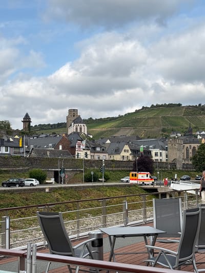 The image captures a view of the town of Oberwesel, Germany, with a focus on its historical architecture and the surrounding landscape. In the foreground, a section of a riverboat's deck is visible, featuring two empty chairs and a small table. Beyond the deck, a stone wall separates the riverbank from a street level. On the street, two cars are parked: a black SUV and a white compact car. Traffic lights are visible, indicating an active road.

Further into the scene, an ambulance with flashing lights is stopped on the side of the road, with two individuals in red vests attending to it. A bridge extends over a lower section of the embankment.

The town itself is characterized by its densely packed buildings with gray tiled roofs. Prominent structures include a tall, dark tower on the left, resembling a watchtower, and a larger building with a bell tower and Gothic architectural elements, possibly a church or cathedral, further to the right. The town is nestled against a steep hillside covered in meticulously maintained vineyards, indicative of the region's wine production. The sky is overcast with scattered clouds. The overall impression is one of a historic European town situated in a scenic, rural setting.