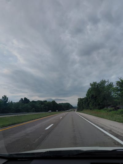 The image captures a view from a moving vehicle on a highway, likely in or near Clay City, United States of America, under a cloudy sky. The highway has multiple lanes with clear white and yellow lane markings. Green trees and foliage line both sides of the road, with some occasional residential structures visible on the right. A few cars are present on the highway, indicating moderate traffic. The overall atmosphere is overcast, suggesting a possibility of rain or an impending storm.