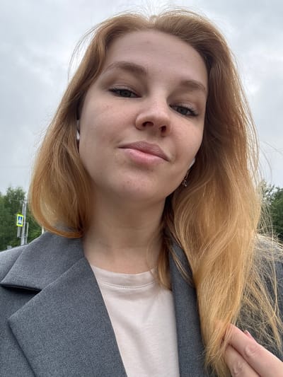 A young woman with shoulder-length, reddish-blonde hair, wearing a grey blazer over a light-colored t-shirt and white earbuds, is captured in a close-up selfie. Her face is angled slightly, and she appears to be outdoors with an overcast sky above. In the background, a green tree line and a yellow pedestrian crossing sign are visible. The setting is indicative of a street scene in the city of Zavolzhye, Russia, likely a suburban or park area given the foliage and pedestrian infrastructure.