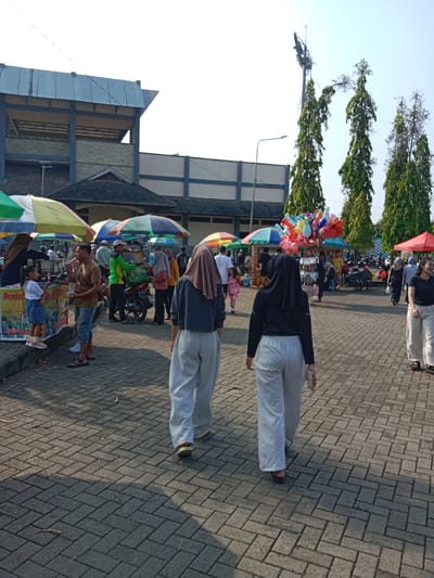 The image depicts an outdoor scene on a paved area, likely a public space in Kendal, Indonesia. Two women are walking away from the viewer, one wearing a grey top and beige hijab with light grey pants, and the other in a black top with a black hijab and light grey pants. The ground is paved with grey brick. In the background, numerous people and vendors are visible under colorful umbrellas, possibly a market or fair. There are food stalls and a balloon vendor among them. To the left, a vendor is seen with a small child, with signage indicating local fare like "Pentol." In the backdrop, there is a building with a grey roof, along with tall trees and a tall lighting structure with bare branches. The lighting and shadows suggest it is daytime.