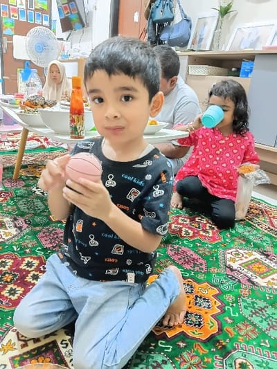The scene depicts an indoor gathering in Meunasahbaet, Indonesia. A young boy, approximately 3-5 years old, kneels barefoot in the foreground on a vibrant green, red, and orange patterned rug. He wears a black t-shirt with white graphics and light blue jeans, holding a small, light pink sphere (resembling a toy basketball) in both hands, looking directly at the camera with a neutral expression. To his right and slightly behind him, a young girl of similar age sits barefoot on the rug, wearing a pink long-sleeved shirt with white hearts and black pants. She holds a light blue plastic cup to her mouth, appearing to drink, with a clear plastic bag of yellow crispy snacks beside her. In the background, a low white table is laden with bowls of food, including pastries and a red-labeled bottle containing orange liquid. Behind the table, a man in a grey t-shirt is partially visible, and to the left, a woman wearing a beige hijab is also partially visible, both seated on the floor. A white pedestal fan stands to the left of the table. The background includes a wall-mounted television, assorted framed pictures, and hanging bags, indicating a residential setting.