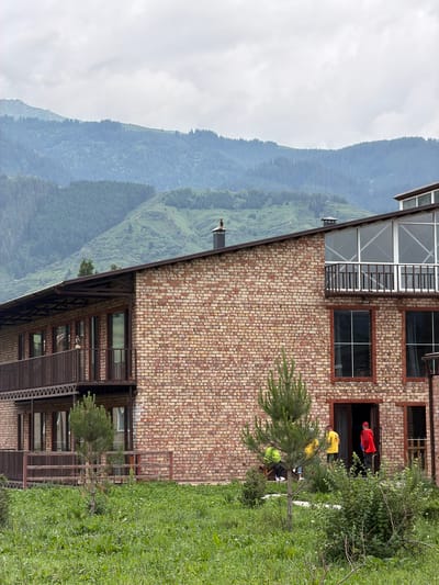 In Shabdan, Kyrgyzstan, a two-story brick building with balconies and large windows stands against a backdrop of verdant, tree-covered mountains under a cloudy sky. The building's brickwork is a mix of yellow and brown tones. The ground floor features an open doorway where three individuals are visible. One person in a red jacket stands outside the doorway, facing away from the camera. Another person in a yellow shirt stands inside the doorway. A third person, also in a yellow shirt, is partially visible near the doorway, with their back to the camera. The area in front of the building is covered in dense, green grass and foliage, with a small pine tree in the foreground. Balconies with brown railings are present on both levels of the building. The mountain landscape is characterized by rolling hills covered in dense evergreen trees, with some patches of lighter green vegetation visible on the slopes. The sky is overcast with gray clouds, suggesting a diffused light and possibly cooler weather.