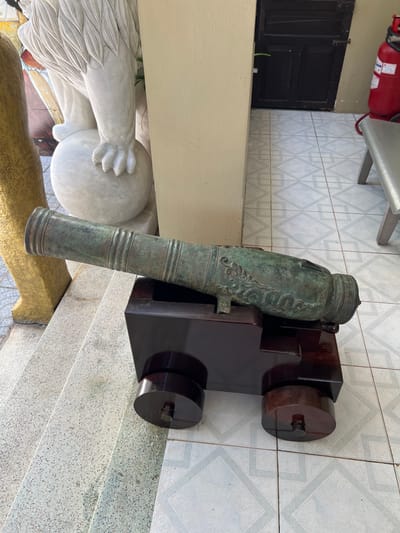 An old cannon rests on a dark wooden carriage with circular wheels. The cannon itself is green, possibly bronze, with ornate designs. It sits beside a marble statue of a figure holding a globe. Behind the cannon, a tan wall with a white-tiled floor is visible. A red fire extinguisher is mounted to the left of the scene, above a black door. The scene is brightly lit.