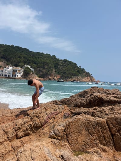 The photo was taken at a rocky beach in Tamariu, Spain, with coordinates 41.918, 3.208. A young man, wearing blue swim trunks and no shirt, is standing on a large, flat rock, looking down at the water below. He is bending over as if he is about to jump into the water. The rock is part of a larger rock formation that extends into the sea, creating a natural barrier between the beach and the open water. The water is a turquoise blue and there are small waves crashing on the shore. In the background, there are a few small boats and a forested hillside with white buildings. The sky is clear and blue, and the sun is shining.