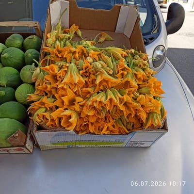 The image shows a close-up view of a box filled with vibrant orange zucchini flowers, likely displayed for sale. To the left, a wooden crate contains several large, smooth green fruits, possibly melons or gourds. The boxes are placed on the hood of a silver vehicle, which appears to be parked on a street. In the background, a building and a parked car are visible, suggesting an outdoor market or roadside stall in a town. The overall scene conveys a sense of fresh produce being offered for sale in an urban or semi-urban setting. The timestamp "06.07.2026 10:17" is superimposed on the image.