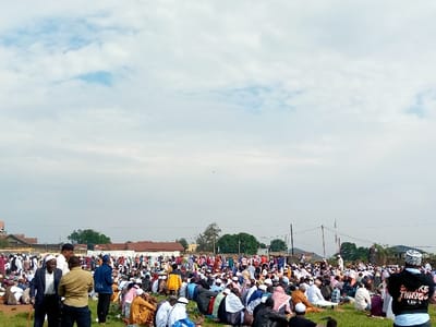 The image shows a large gathering of people, possibly a religious assembly or a public event, taking place on a grassy field in Beni, Democratic Republic of Congo. The location is likely a public space, possibly a park or a field adjacent to a mosque, as indicated by the presence of the people and the general setting.  There are over 100 people present, mostly men, wearing various clothing, some with traditional white headwear, others with casual attire, and a few with a hoodie and jeans. The attendees are seated on the ground, engaging in conversation and prayer. The field is mostly covered with green grass with a few visible trees in the background, indicating an outdoor setting. The sky is cloudy with some sunlight. It appears to be a sunny day in the afternoon or evening.