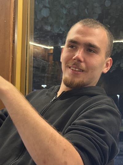 A young man with short hair and a goatee is seated indoors, with his arm resting forward. He is wearing a black zip-up hoodie. Behind him, a window reflects the outdoors at night, with some faint lights visible. The scene appears to be from Lom, Bulgaria.