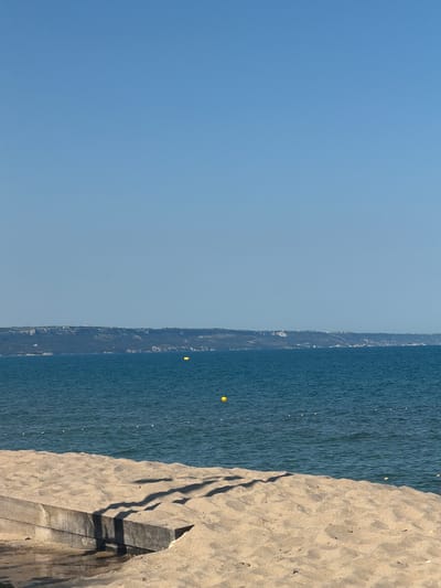 The image depicts a serene beach scene in Kranevo, Bulgaria. The foreground is dominated by a sandy beach with a concrete structure, possibly a retaining wall or a part of an old pier, partially covered in sand. Shadows from an unseen object, likely a tree or a building, are cast across the sand and the concrete.

Beyond the beach, the deep blue sea stretches towards a distant coastline. The water is calm, with gentle ripples and a few small white buoys marking an area. In the mid-ground, a few yellow buoys are visible, and a thin rope with white markers stretches across the water, possibly indicating a swimming zone or a boundary.

The distant coastline features a range of hills or cliffs covered in vegetation, with a few scattered white structures visible on top, suggesting a town or settlement. The sky is a clear, cloudless blue, indicating a sunny day. The overall impression is one of a quiet, peaceful day at the seaside in Kranevo, Bulgaria.
