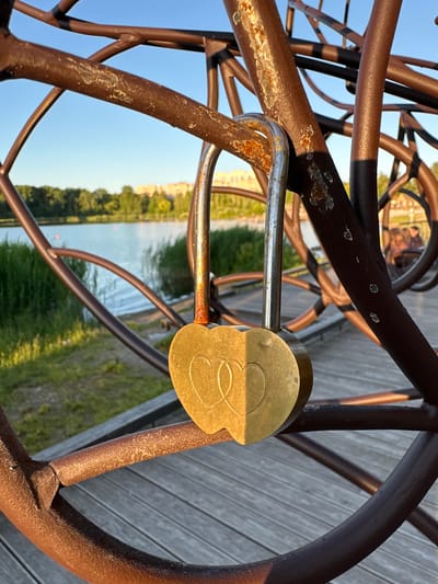 A close-up shot focuses on a heart-shaped padlock attached to a weathered, abstract metal sculpture in Solnechnogorsk, Russia. The sculpture, made of thick, rusty metal bars, forms a circular pattern with a dark, almost black, finish in some areas and a reddish-brown, distressed texture in others. The padlock is brass, with two interlocking heart outlines etched into its surface. It is secured to one of the sculpture's bars, which shows signs of rust and wear.
In the background, a body of water, likely a lake, stretches across the frame, bordered by tall green reeds and a grassy embankment. Beyond the water, a line of trees with lush green foliage is visible, and in the far distance, several light-colored buildings can be seen, suggesting an urban or suburban setting. The sky above is a clear, bright blue, indicating a sunny day. A wooden boardwalk or pier is present beneath the sculpture, with visible wooden planks. The overall scene evokes a sense of romantic tradition, with the padlock symbolizing a bond of love, set against a tranquil natural backdrop within the city.