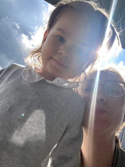 An outdoor selfie features two individuals, a young child and an adult, captured at a low angle against a bright sky in Kharnavy, Russia. The child, a young girl with brown hair, is in the foreground, looking directly at the camera with a neutral expression. She is wearing a grey sweater with a distinctive white, lacy Peter Pan-style collar.
Behind and slightly to the right of the child, an adult, likely a woman, is partially visible. She wears glasses and also looks towards the camera. Both individuals appear to be wearing grey-toned tops.
The image is characterized by strong backlighting from the sun, which is positioned in the upper left, causing prominent lens flare and bright rays of light to stretch across the frame. This intense sunlight creates a halo effect around the subjects' hair and illuminates hair strands, indicating daytime conditions. The background shows a bright blue sky with some white clouds. No specific objects or visible text are present, apart from the subjects and their attire. The overall impression is a casual, sun-drenched moment captured outdoors.