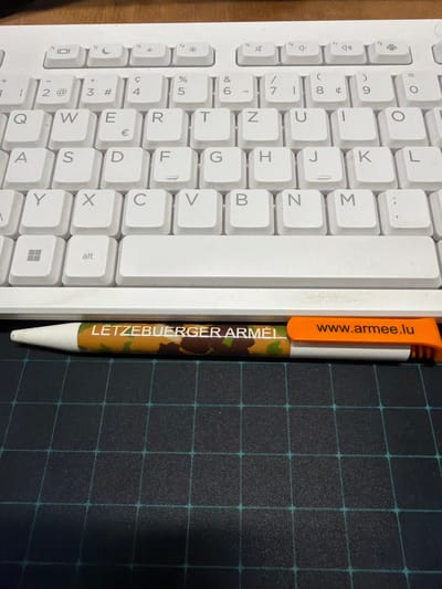 A top-down view shows a section of a white keyboard adjacent to a pen and a black-squared mousepad. The keyboard features standard alphanumeric keys and a blank white bar. The pen has an orange clip with the text "www.armee.lu" and a camouflage pattern with "LËTZEBUERGER ARMÉI" vertically printed on its side. The mousepad is dark with a grid pattern of light blue lines. There is no information to suggest a specific location in Pétange, Luxembourg.