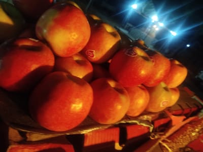The image depicts a close-up, low-angle shot of a pile of red apples. The apples are arranged in a woven basket or crate, with some appearing to have small white stickers on them. The lighting is dim and appears to be from overhead bulbs, creating highlights and shadows on the fruit. The background is dark and out of focus, with some blurry lights visible, suggesting an indoor or nighttime setting. The context of Jimeta, Nigeria is not visually present in the image itself.