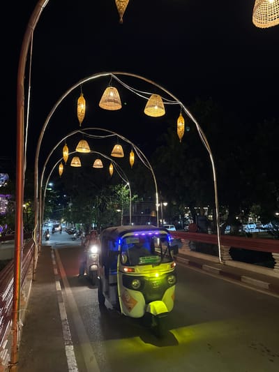This photograph captures a nighttime scene on a bridge in Siem Reap, Cambodia, likely part of the Pub Street area or a connecting thoroughfare. The bridge is adorned with a series of large, arched metal structures, each lined with white fairy lights. Suspended from these arches are numerous lamps, predominantly woven from natural materials with a conical or bell shape, emitting a warm, golden light. The street below is paved and shows signs of traffic. In the foreground, a bright yellow auto-rickshaw, also known as a tuk-tuk, is prominently featured, its headlights illuminating the road ahead and casting a yellow glow. A couple is visible riding a scooter towards the tuk-tuk, their faces partially obscured by helmets. The background reveals a street with other vehicles, including cars and motorcycles, and the silhouettes of trees and buildings. The overall atmosphere is festive and illuminated, suggesting a lively urban environment at night.