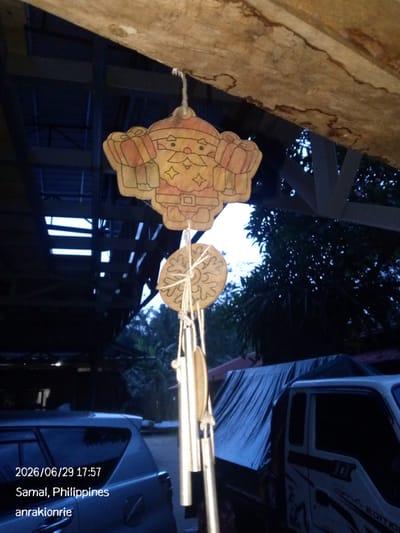 A close-up, low-angle, outdoor shot captures a wooden wind chime hanging from a wooden beam. The wind chime features a Santa Claus-like figure at the top, followed by a circular snowflake ornament, and several metallic tubes. The background is dark, with trees and part of a structure with visible beams and openings suggesting a roof or shelter. A blue tarp covers the bed of a white truck parked to the right, and a portion of a silver SUV is visible on the left. A timestamp and location are overlaid on the image: "2026/06/29 17:57 Samal, Philippines anrakionrie."