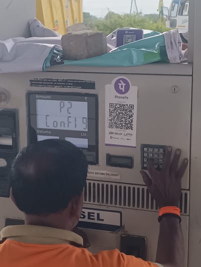 The image shows a man operating a fuel dispenser. The dispenser has a digital display showing "P2" and "Conf19". There is a QR code for PhonePe and BHIM UPI on the dispenser. The man is wearing an orange shirt and an orange wristband. In the background, there is a yellow truck and greenery. The location is specified as Sugalimetta, India.