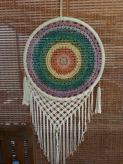A macrame wall hanging is centered against a bamboo-slatted wall. The wall hanging features a circular crocheted design with concentric rings of various colors, resembling a rainbow. The outer edge of the circle is wrapped in white cord. From the bottom of the circle, white macrame cords hang, adorned with small wooden beads. Two tasseled white cords hang from the sides of the circle, also with wooden beads. To the left of the wall hanging, the corner of a dark wooden chair is visible. The scene appears to be indoors, with the bamboo wall suggesting a rustic or bohemian decor style.