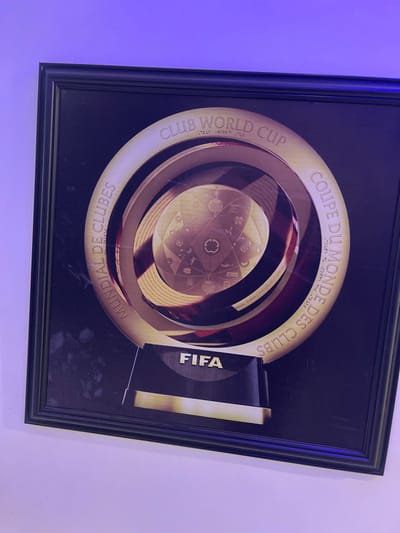 A framed picture depicting the FIFA Club World Cup trophy is mounted on a light-colored wall illuminated by a purple light source. The image, encased in a black frame, features a gold and dark-toned representation of the trophy. Inscriptions around the circular trophy design read "CLUB WORLD CUP," "MUNDIAL DE CLUBES," and "COUPE DU MONDE DES CLUBS." The base of the trophy image prominently displays "FIFA." Faint smudges are visible on the left side of the print within the frame. No individuals or active interactions are present. This scene is located in Orie Uratta, Nigeria.
