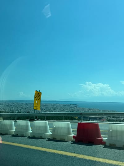 The image, taken from a vehicle, shows a view overlooking the city of Agios Pavlos Municipal Unit, Greece. In the foreground, a row of white and one red plastic traffic barriers are lined up along the side of a road with yellow lane markings. A yellow road sign with three black arrows pointing diagonally upwards and the text "200m" is visible behind the barriers. Beyond the road, the dense urban landscape of Agios Pavlos Municipal Unit stretches towards the sea. The sea itself is visible, with landmasses and distant mountains on the horizon. The sky is clear and blue with a few scattered white clouds. The scene depicts a roadside view with infrastructure and a panoramic vista of a coastal Greek city.