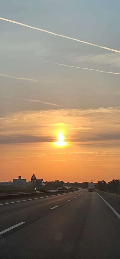 The image depicts a sunset view from a highway likely in or near Balatonfőkajár, Hungary. The sun, a bright orb, is setting low in the sky, casting a warm orange glow and illuminating thin, elongated clouds. Several faint contrails from aircraft are visible in the pale blue sky above.

The highway itself is a multi-lane road with clear lane markings. In the foreground, the road surface is visible, with shadows cast by unseen objects. In the distance, several vehicles are present on the highway, including what appears to be a truck in the right lane. On the left side of the highway, there is a large, boxy structure that might be a building or industrial facility, and a triangular road sign. Trees and vegetation line the sides of the highway, forming a natural backdrop to the scene. The overall impression is one of a tranquil evening drive.