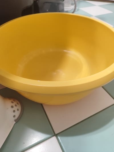 A yellow plastic bowl is centered in the frame, occupying most of the image. The bowl has a smooth, rounded interior and a raised rim. The exterior is not fully visible, but a portion of its curve is apparent. The bowl rests on a tiled surface. The tiles are arranged in a diagonal grid pattern and appear to be a light green and off-white color. In the lower left corner of the image, a portion of a white object with grey circular perforations is visible, partially obscured by the yellow bowl. In the background, out of focus, there is a dark object and hints of a light-colored surface. The lighting appears to be overhead, casting soft shadows on the bowl and tiles.