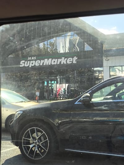 A daytime external scene depicts the frontage of an "İnkas SuperMarket" building in Kızılbaş, Northern Cyprus / Cyprus. The building features a dark facade with expansive reflective glass windows, including a prominent triangular section on the upper level, and a dark, possibly tiled, roof. A black Mercedes-Benz GLC SUV is parked in the foreground, partially visible on the right, with its front passenger wheel, fender, and side mirror clearly in view. To its left, a gold/beige sedan is parked, reflecting strong sunlight on its windshield.
In the mid-ground, one individual, dressed in a light-colored top and white shorts, is observed from behind, walking towards the supermarket entrance. A rack holding multiple bicycles, mostly dark in color, is positioned near the entrance area. Reflections of trees and the sky are visible in the supermarket's glass facade. A white sign with red text or graphics is visible near the entrance. The ground surface in the foreground appears to be paved, indicative of a parking area. The sky is clear blue with minimal cloud cover, and bright sunlight is present, causing glare on the vehicles and the apparent vantage point of the photographer. A security camera is mounted on the lighter-colored building section to the right of the main supermarket structure.