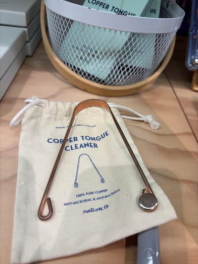 The image shows a copper tongue cleaner lying on top of its cloth storage bag, which is placed on a wooden surface. The bag has blue text that reads "COPPER TONGUE CLEANER" and other descriptive phrases. The tongue cleaner is a U-shaped metal instrument, with loops at either end. In the background, a white mesh basket with a wooden rim contains what appears to be a book or pamphlet. The scene is brightly lit, with a shallow depth of field that blurs the background elements. The location in the city of Antigua, Spain, is not discernible from the provided image.