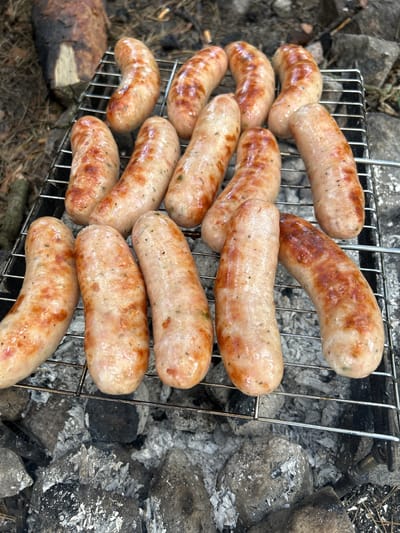 The image depicts a close-up view of numerous sausages being grilled on a metal rack over hot coals. The sausages appear to be fully cooked, with a golden-brown exterior and some char marks. The grilling rack is positioned over a bed of grey and white ashes, interspersed with larger rocks. In the background, out of focus, a piece of wood is visible, possibly part of a campfire. The overall setting suggests an outdoor, possibly rustic environment, consistent with a picnic or camping scenario. The image does not provide any information about the specific location in Chmyrivka, Ukraine, or the presence of any people.