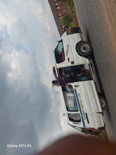 A white passenger van, identified by "ENTRACO" on the front door and "ENUGIRT COMPANY LTD" on its side, is stopped on an asphalt road in Awkuzu, Nigeria. The vehicle's passenger-side sliding door is open, revealing multiple occupants inside. One individual, wearing a striped garment with red, blue, and white bands over a dark shirt, stands outside the open door, facing into the van, indicative of boarding, alighting, or interaction with passengers. The van shows signs of operational wear. The environment consists of a dark asphalt road surface, a dirt shoulder with sparse green vegetation to the vehicle's right, and a slightly elevated background feature possibly incorporating a concrete or earth embankment with visible metallic elements. The sky above is partly cloudy.