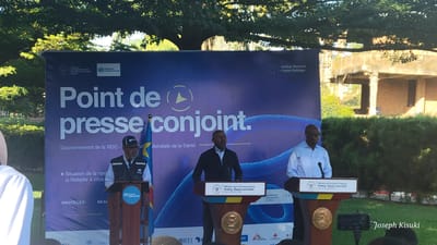 The image captures a joint press conference in Bunia, Democratic Republic of the Congo, likely related to public health concerns. Three men stand behind podiums, addressing an unseen audience. The backdrop is a large banner titled "Point de presse conjoint" (Joint Press Conference) with logos of the World Health Organization and the Institut National de Santé Publique (National Institute of Public Health). A flag of the Democratic Republic of the Congo is visible behind the man on the left. The podiums are branded with the ministry logos and a briefing title related to the Ebola response. A camera is positioned to record the event. Behind the banner, there is greenery and a partially constructed building. A watermark with the name "Joseph Kisuki" is visible at the bottom right.